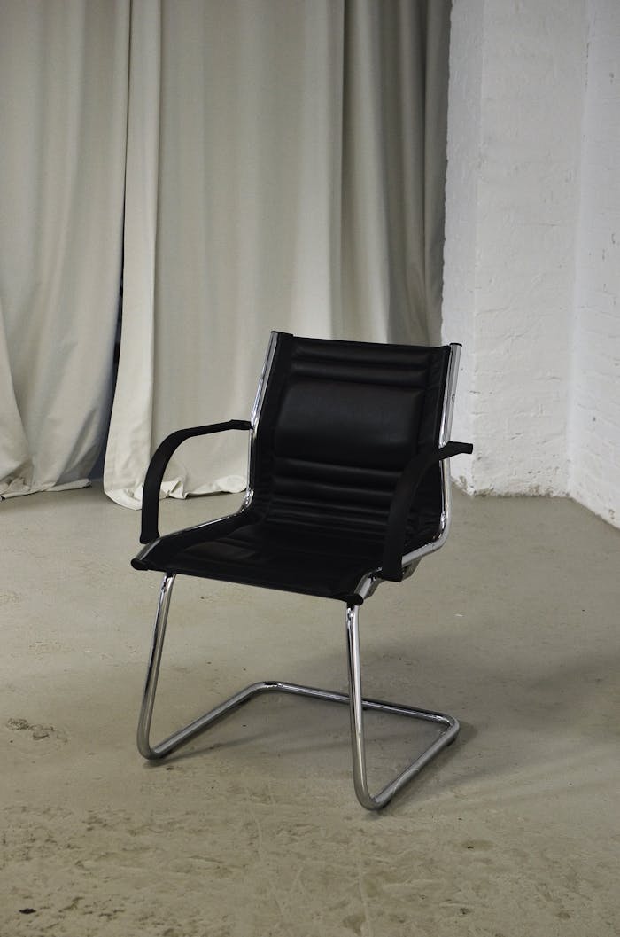 Comfortable office chair with black leather seat and metal elements placed in room with light brick wall and curtain